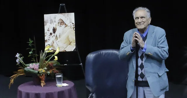 Ishwar Puri speaking on a stage beside flowers and a portrait